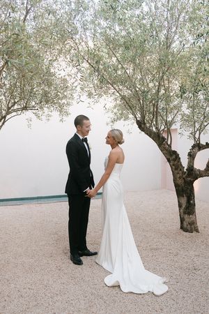 Bride and groom in elegant wedding attire stand holding hands under olive trees on a gravel courtyard.