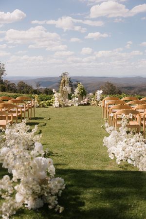 Outdoor wedding ceremony setup with white floral aisle, wooden chairs, floral arch, and panoramic mountain views under blue sky.