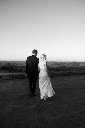 Black and white photo of a bride and groom walking hand in hand across a lawn with scenic countryside views.