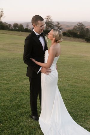 Bride and groom embrace on green hillside at sunset, she wears strapless white gown, he wears black tuxedo with bow tie.