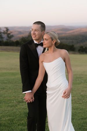 Bride and groom holding hands at sunset on a green hillside, she in a strapless white gown, he in a black tuxedo.