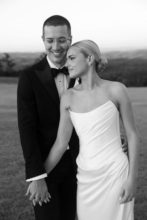 Black and white photo of smiling groom in tuxedo holding bride in strapless wedding gown outdoors.