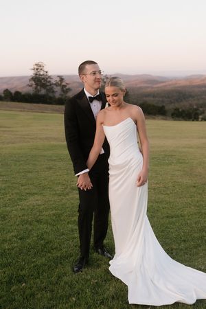 Bride in strapless white gown and groom in black tuxedo hold hands on green hillside at sunset with mountain views.