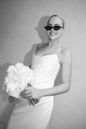 Smiling bride in strapless white gown and cat-eye sunglasses holding a white floral bouquet, black and white photo.