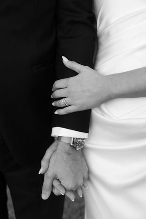 Black and white close-up of bride and groom holding hands, showcasing her diamond ring and his luxury wristwatch.
