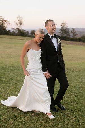 A bride in a white strapless gown and groom in a black tuxedo walk hand in hand on a green lawn at sunset.