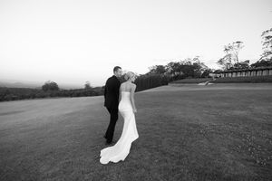 Black and white photo of a bride and groom sharing a romantic kiss on a lush green lawn at dusk.