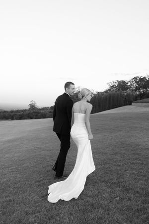 Black and white photo of a bride and groom standing on a grassy hill at dusk, sharing a romantic moment.