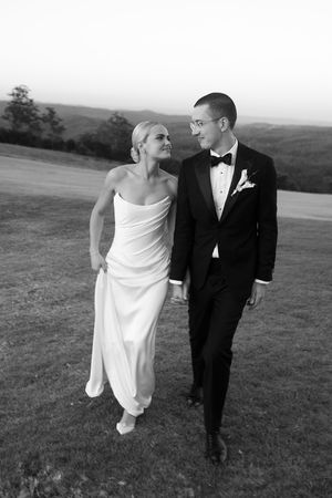Black and white photo of a bride and groom walking together on a grassy hill, smiling at each other on their wedding day.