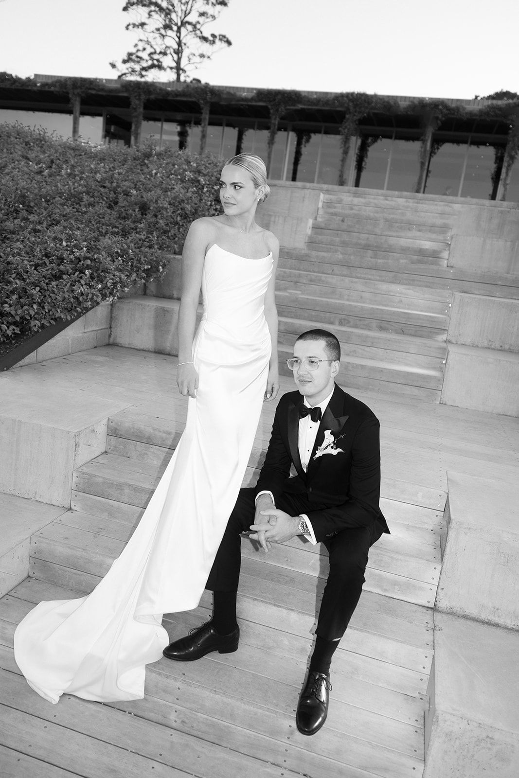 Black and white wedding photo of bride in white gown and groom in tuxedo posing on stone steps outdoors.