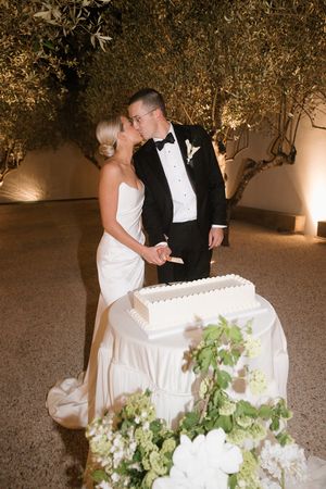 Bride and groom kissing while cutting white wedding cake outdoors at night, surrounded by olive trees and floral arrangements.