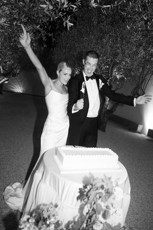 Joyful newlyweds celebrate in black and white photo beside white wedding cake outdoors amid olive trees.