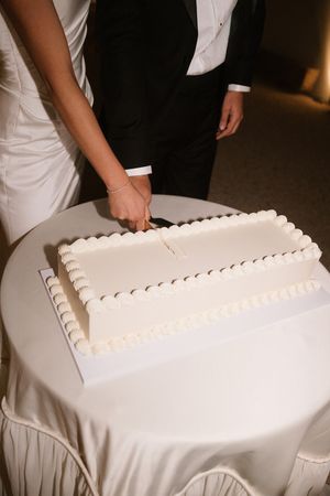 Bride and groom cutting a white rectangular wedding cake with pearl border decoration on a draped table.