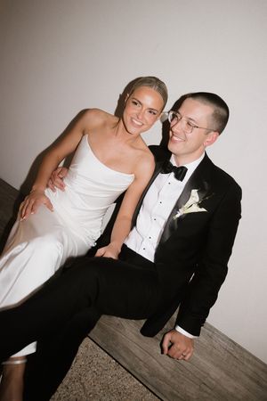 Couple in wedding attire sitting together, bride in white gown and groom in black tuxedo with boutonniere, smiling.