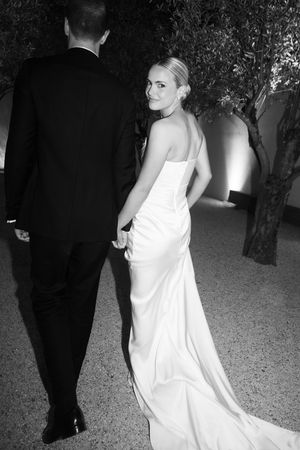 Bride in strapless white gown holds grooms hand walking among olive trees in black and white photo.