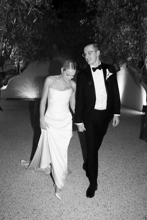 Black and white photo of a bride and groom holding hands walking outdoors at night, she in a strapless gown, he in a tuxedo.