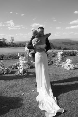 Black and white photo of a bride in a white gown embracing a child outdoors at a wedding ceremony.