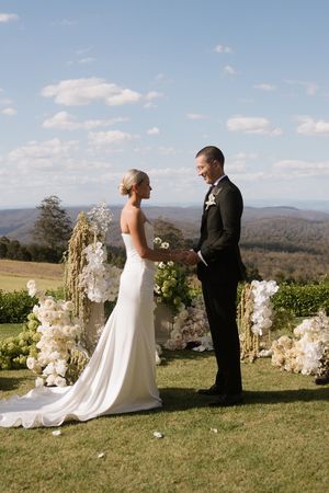 Bride and groom exchange vows outdoors with white floral arch and scenic mountain valley views on a sunny wedding day.