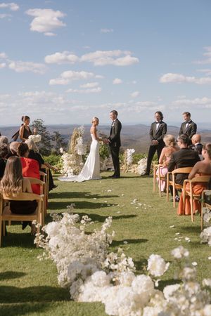 Outdoor wedding ceremony on a hilltop lawn with white floral aisle, guests seated, bride and groom at altar under blue sky.