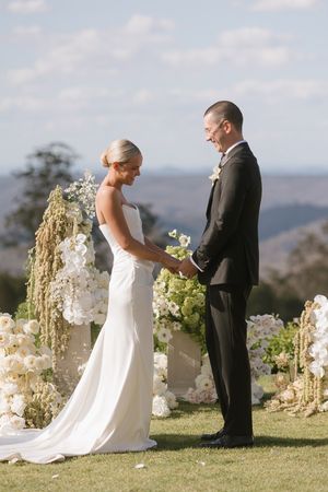 Bride and groom hold hands at outdoor wedding ceremony surrounded by white floral arrangements with scenic hills backdrop.