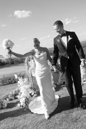 Black and white photo of a bride in white gown and groom in tuxedo walking hand in hand after their outdoor wedding ceremony.