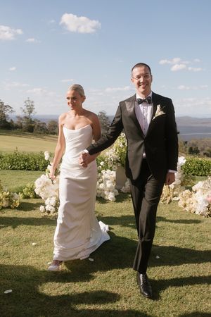A smiling bride and groom walk hand in hand across a lush green lawn with scenic views and white floral arrangements.
