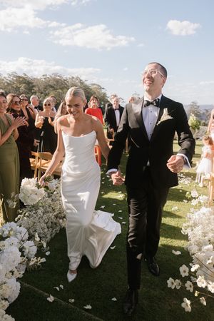 Bride and groom walk joyfully down outdoor aisle lined with white flowers after wedding ceremony.