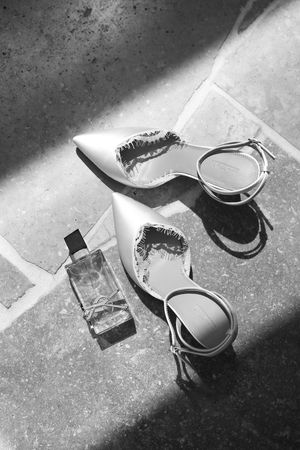 Black and white photo of elegant pointed-toe heels with ankle straps beside a perfume bottle on stone floor.