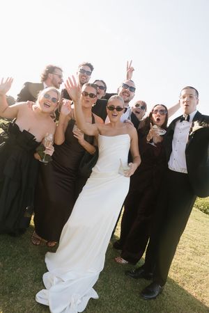 Bride in white gown poses with wedding party in black attire, wearing sunglasses and celebrating outdoors on sunny day.