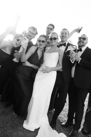 Black and white wedding photo of bride in white gown with bridal party in formal attire, all wearing sunglasses and celebrating.
