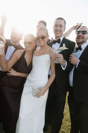 Bride in white gown with wedding party in black tuxedos and dresses celebrating outdoors with drinks in sunlight.