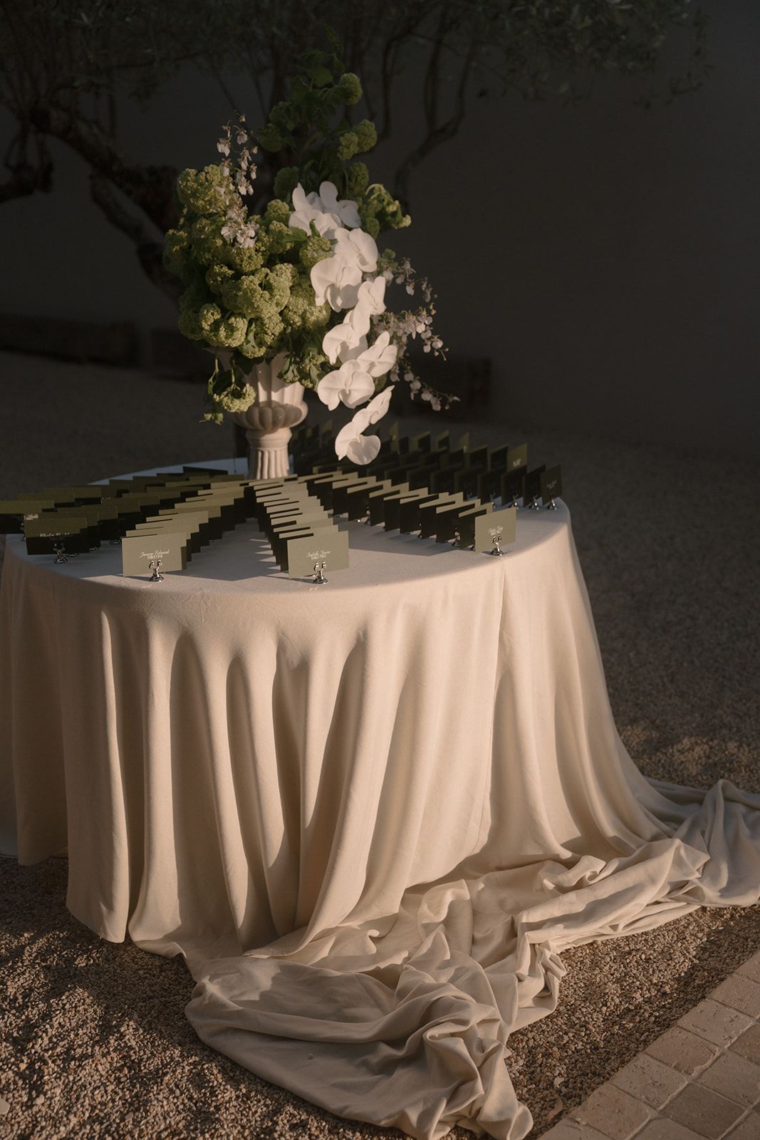 Elegant wedding escort card table with cream linen, place cards, and white orchid floral arrangement outdoors at dusk.
