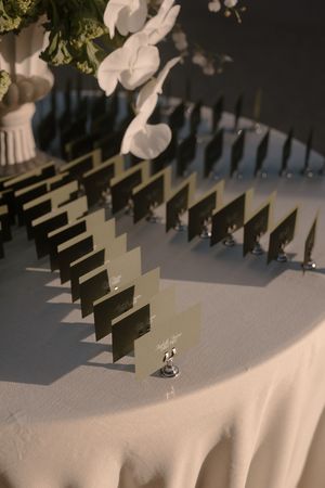 Elegant wedding escort cards displayed in silver holders on a white tablecloth with white orchid floral arrangement.