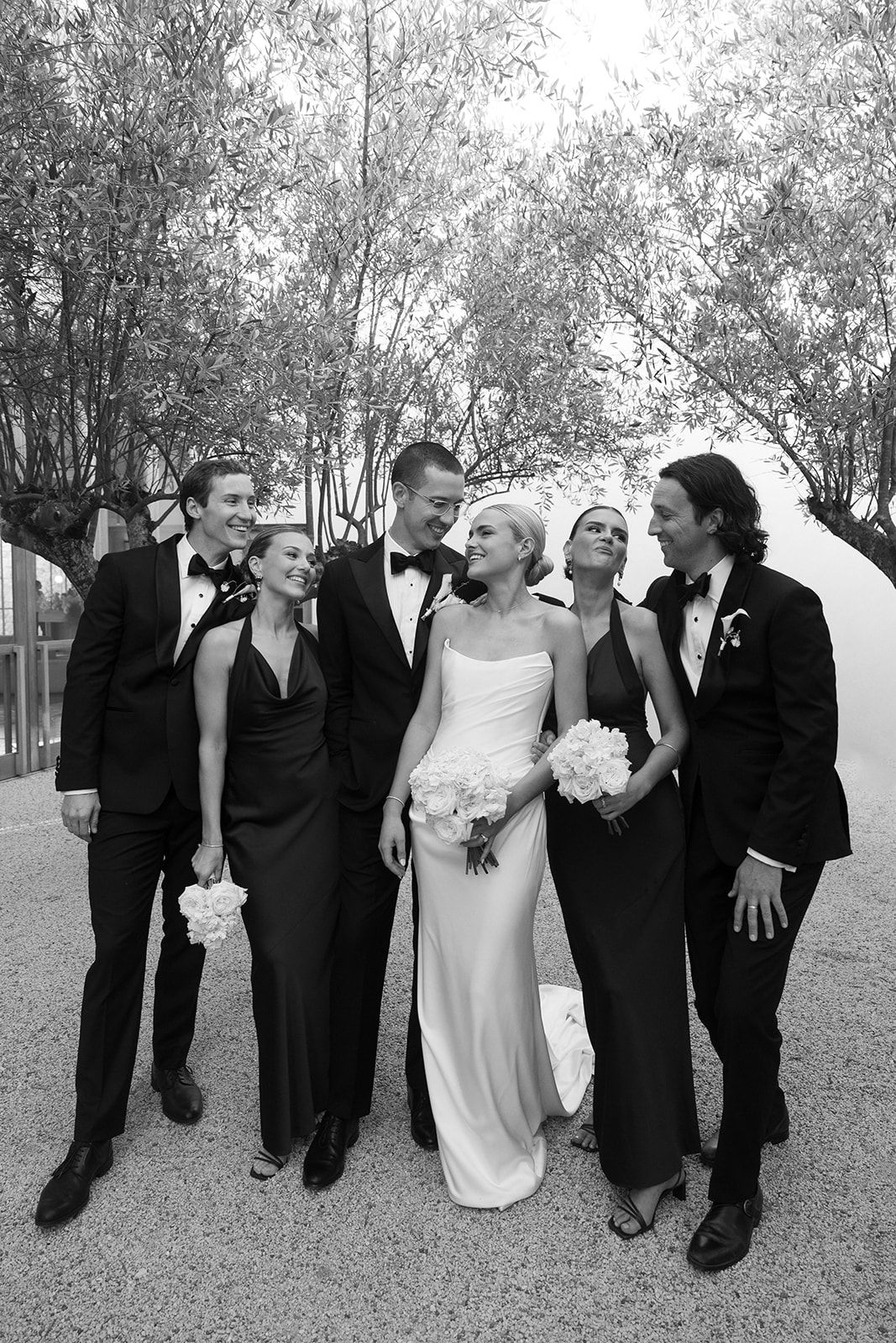 Black and white wedding photo of bride in white gown with bouquet, groom in tuxedo, and wedding party in formal attire.