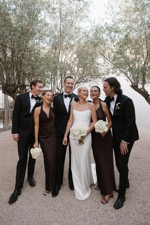 Wedding party in black tuxedos and brown gowns laughing outdoors with bride in white strapless dress holding white bouquet.