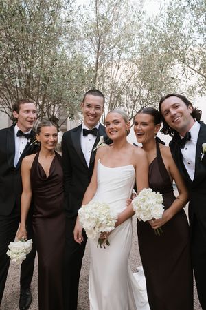 Wedding party posing outdoors, bride in white gown with bouquet, bridesmaids in brown dresses, groomsmen in black tuxedos.