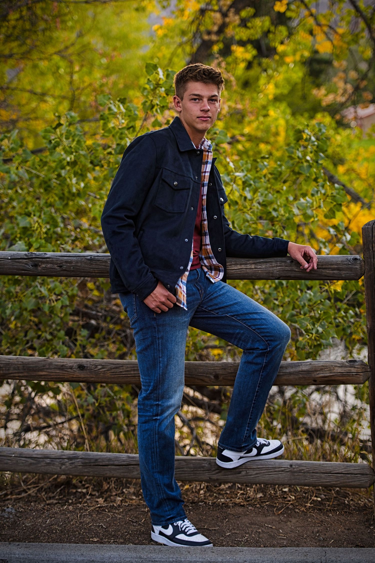 Young man in navy jacket and jeans leaning on rustic wooden fence with autumn foliage backdrop.