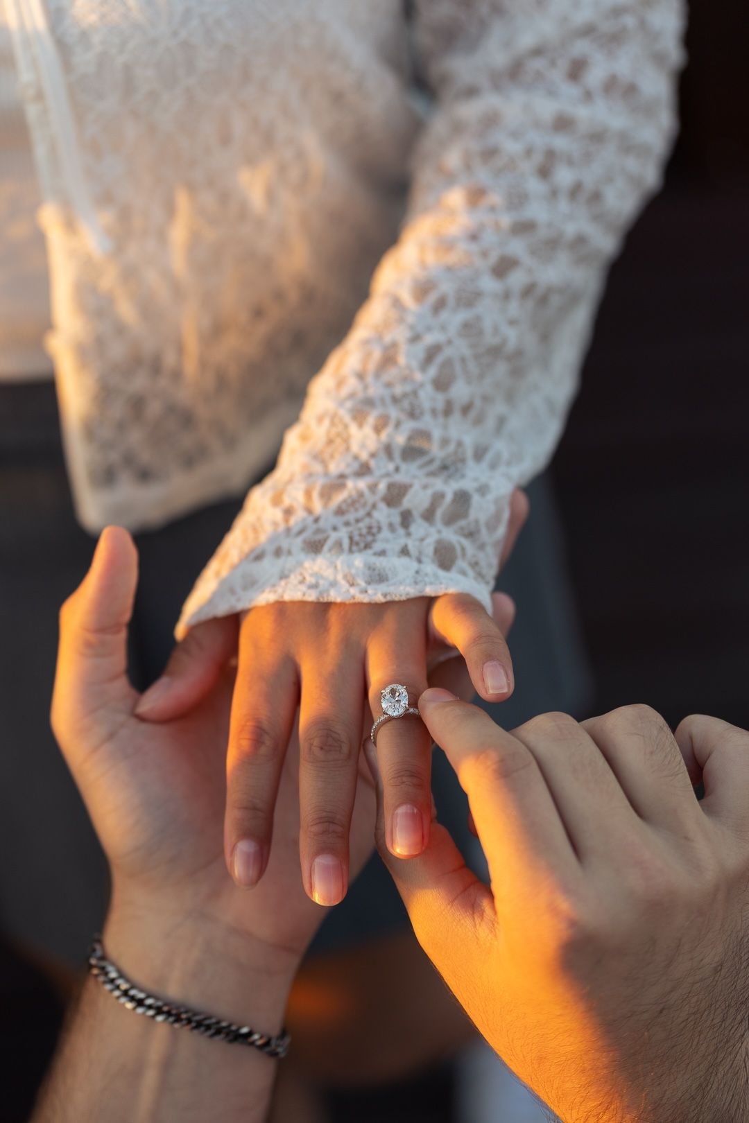 Man placing a diamond engagement ring on a woman's finger, wearing white lace sleeve, in warm golden light.