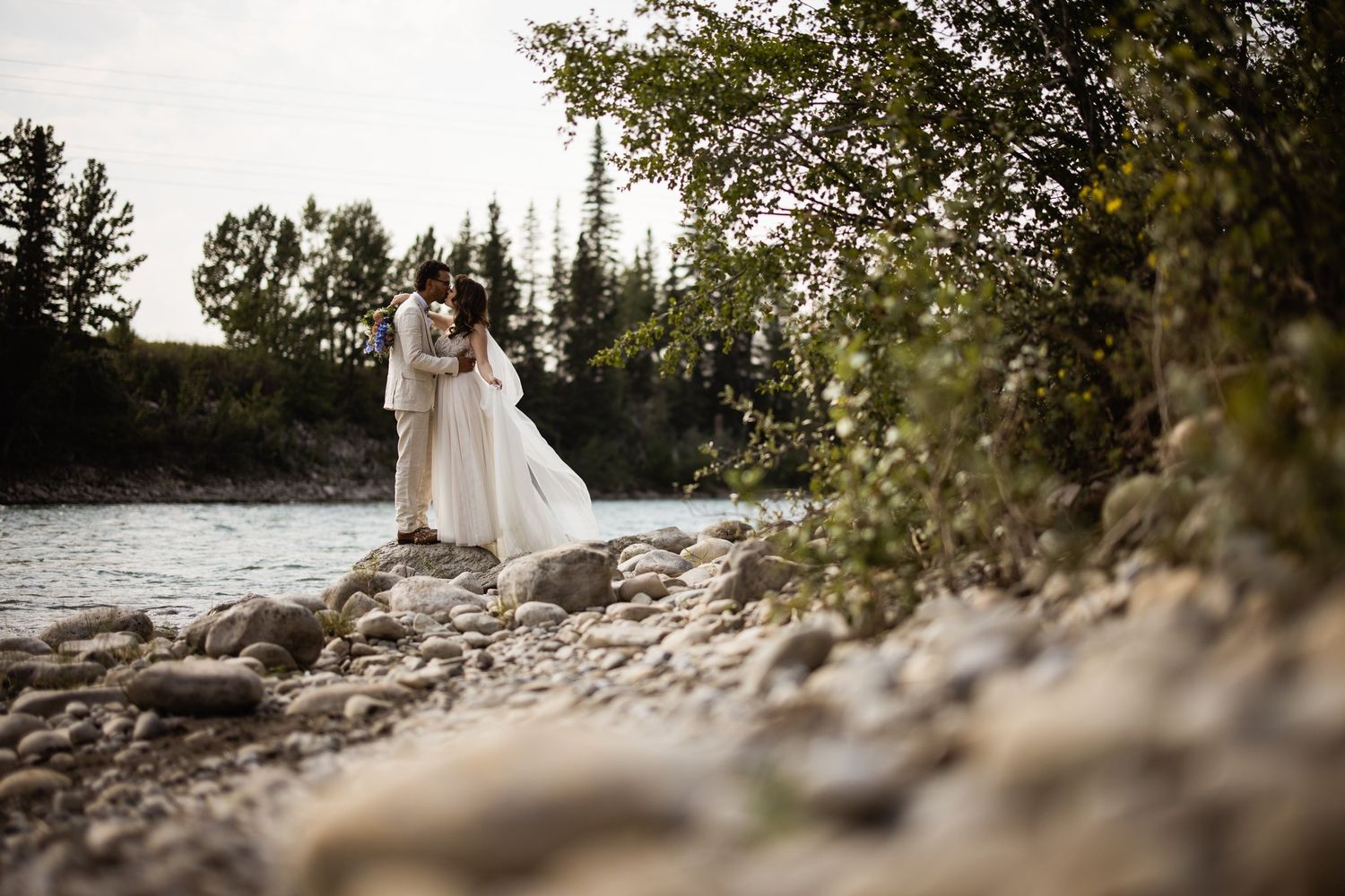 Bride and groom embracing on river rocks with evergreens behind