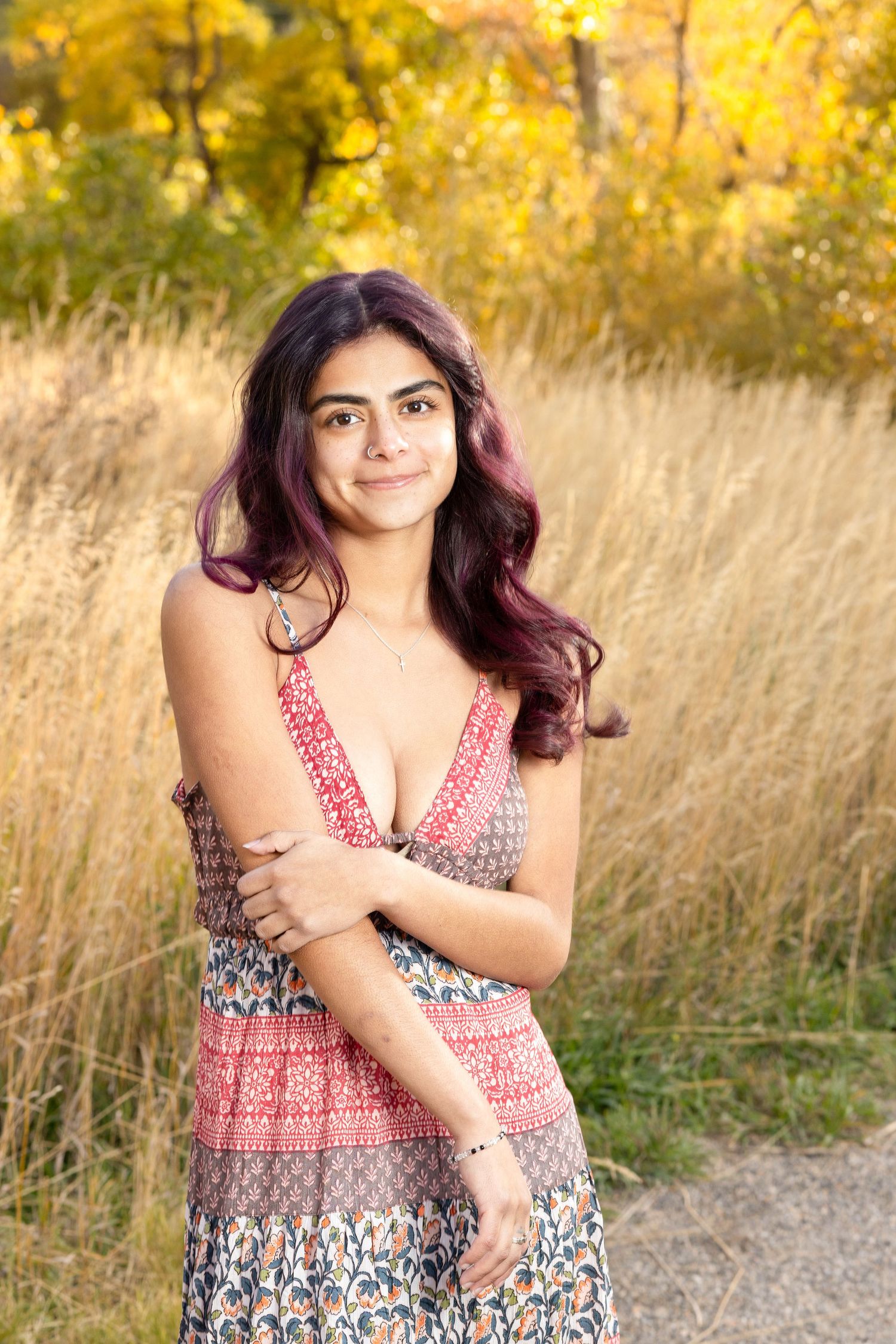 Young woman with purple-tinted hair in a floral dress poses outdoors among golden autumn grasses.