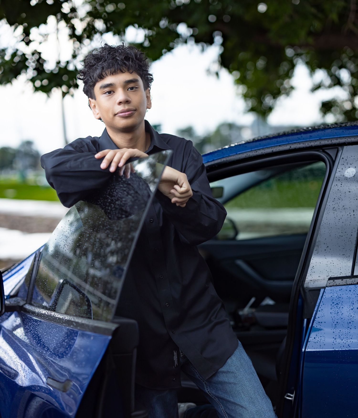 Young man in black shirt leaning on open car door of blue vehicle outdoors.