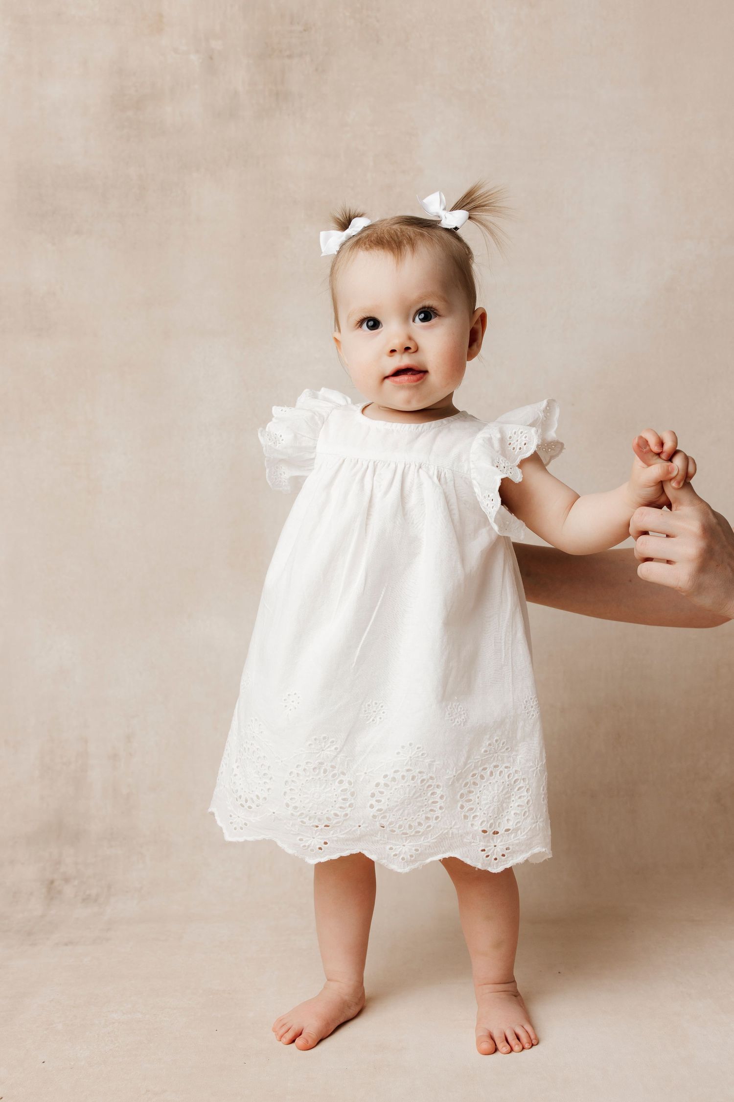 Baby girl in white dress sits holding a sage green number 1 balloon for her first birthday portrait.