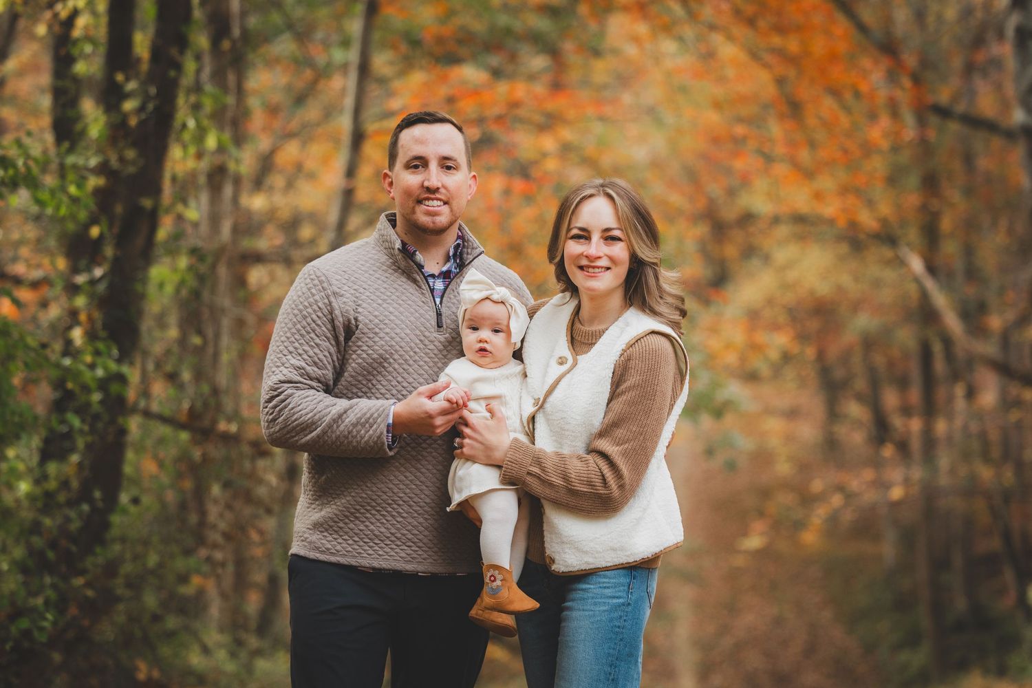 Family of three posing outdoors in autumn forest with colorful fall foliage, parents holding baby in white outfit. at Lake Fairfax Park