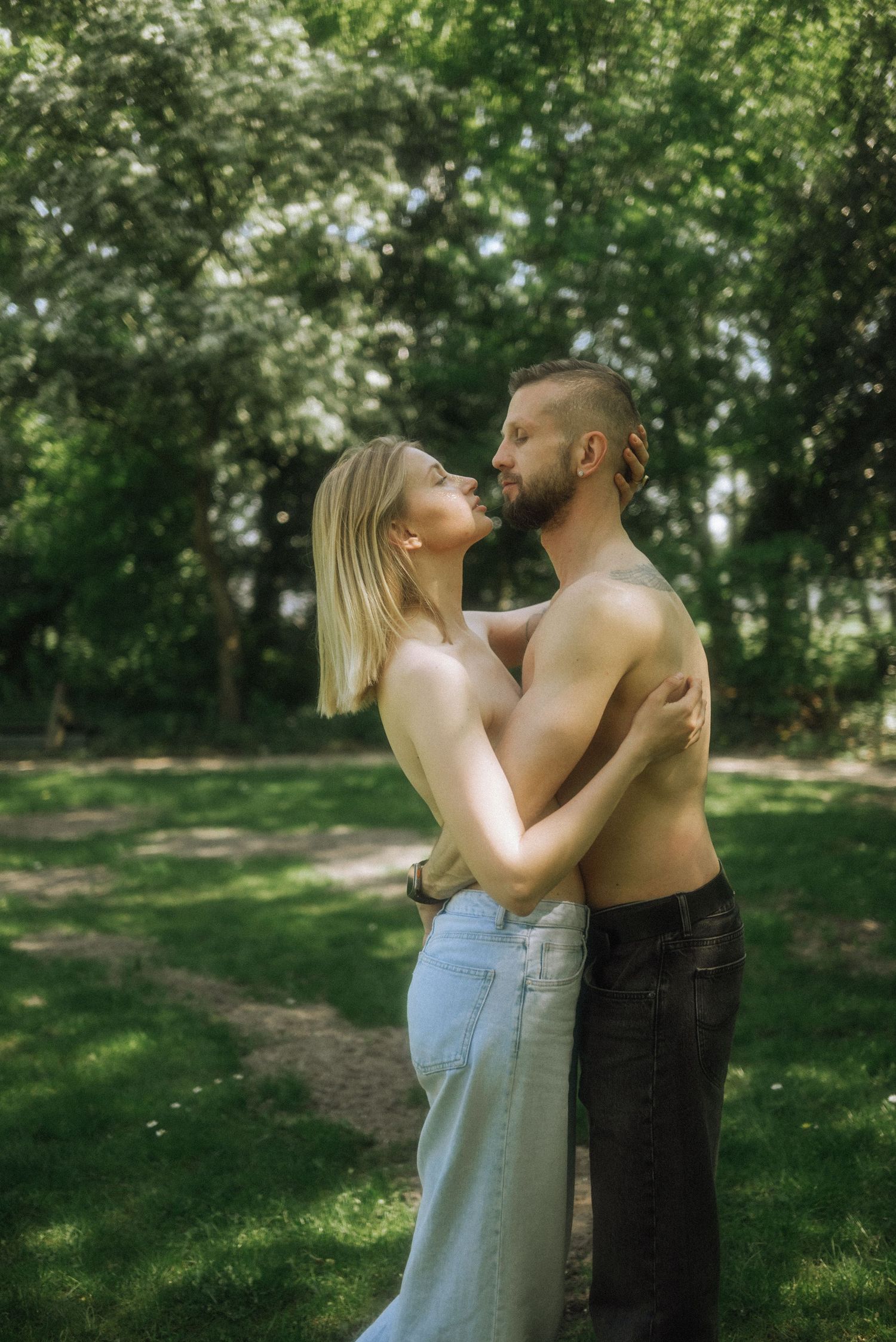 Shirtless bearded man and blonde woman in light jeans embrace face to face in a sunlit green park, Lisse, Huys Dever.
