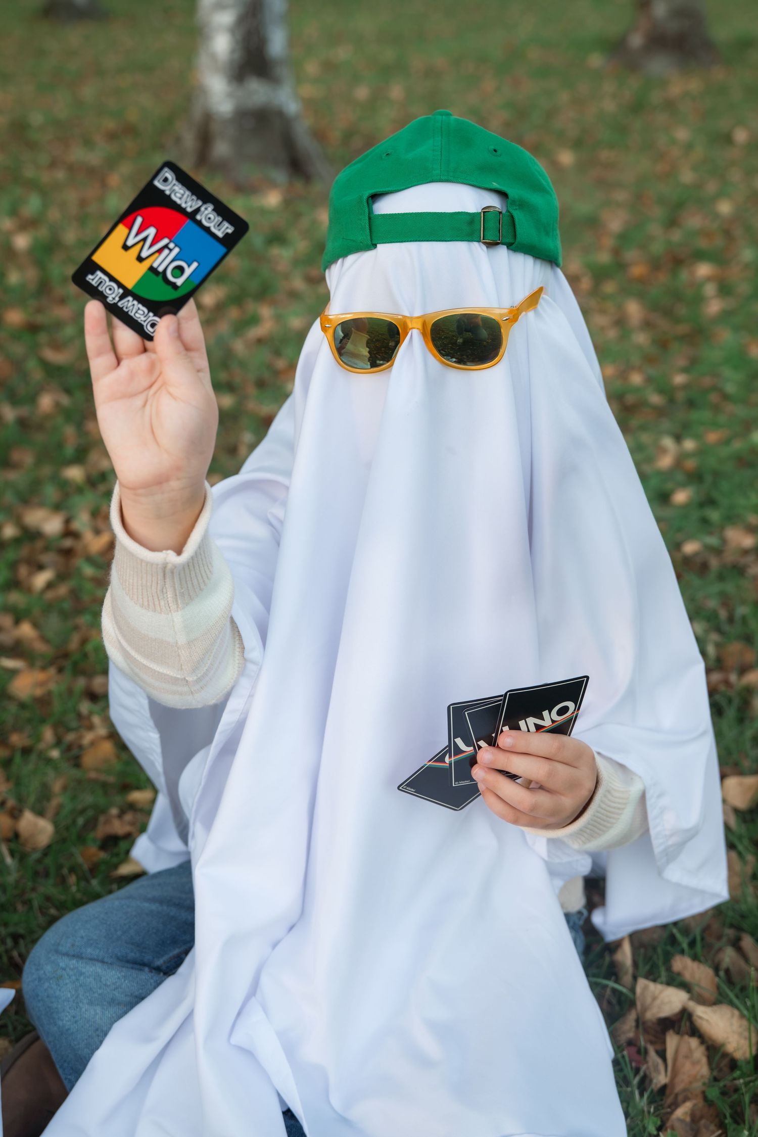 Person in a white ghost sheet, green cap and orange sunglasses holds a Wild Draw Four UNO card while playing UNO outdoors on autumn leaves.