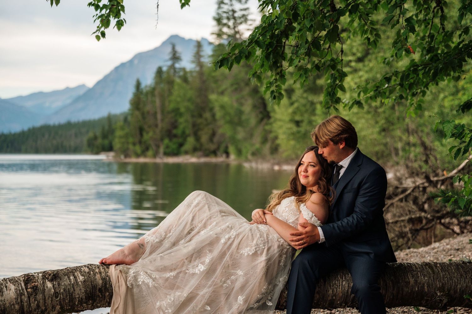 Couple in wedding attire sitting on a log by a mountain lake surrounded by lush forest.