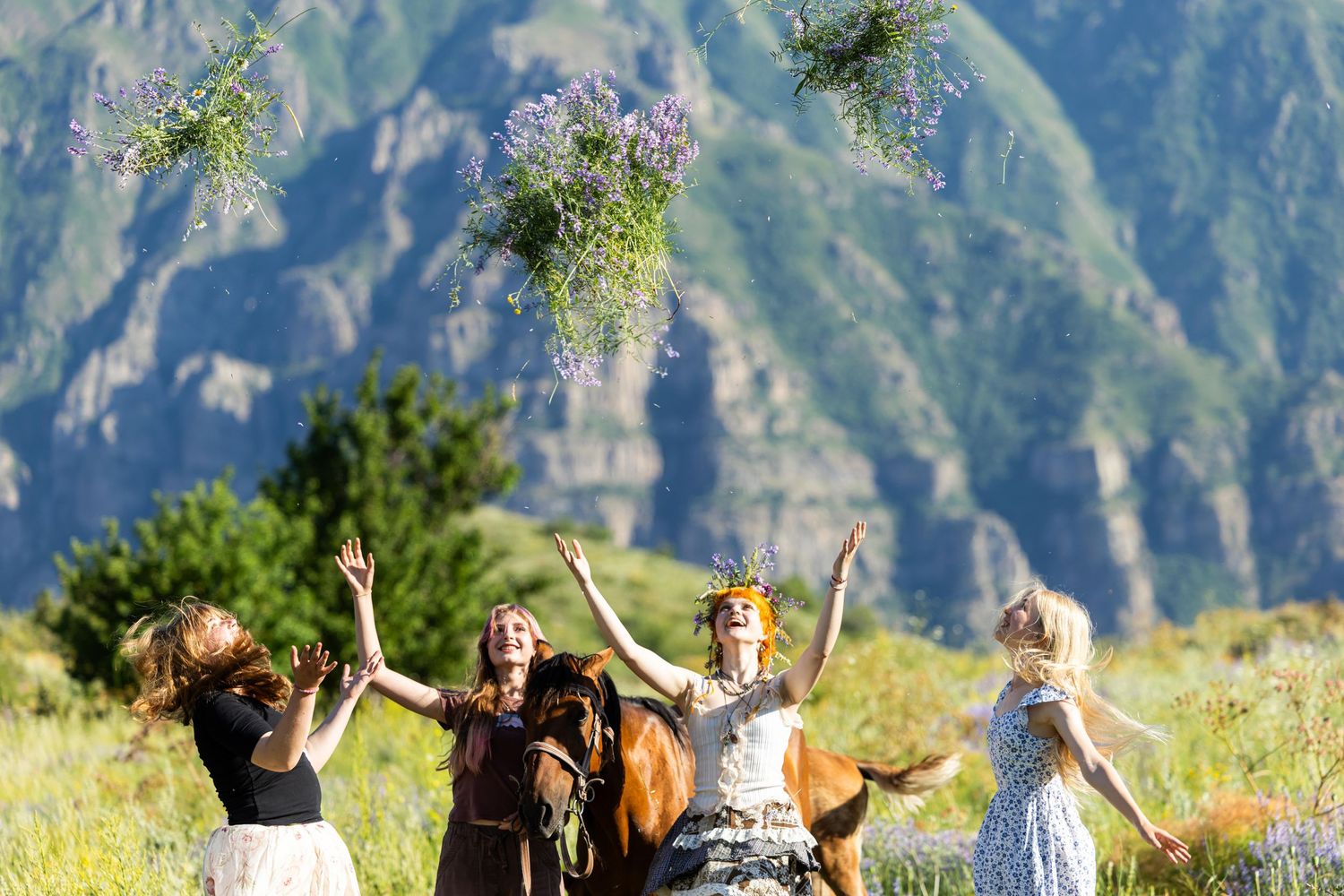 Four women joyfully tossing wildflower bouquets into the air with a horse in a mountain meadow.