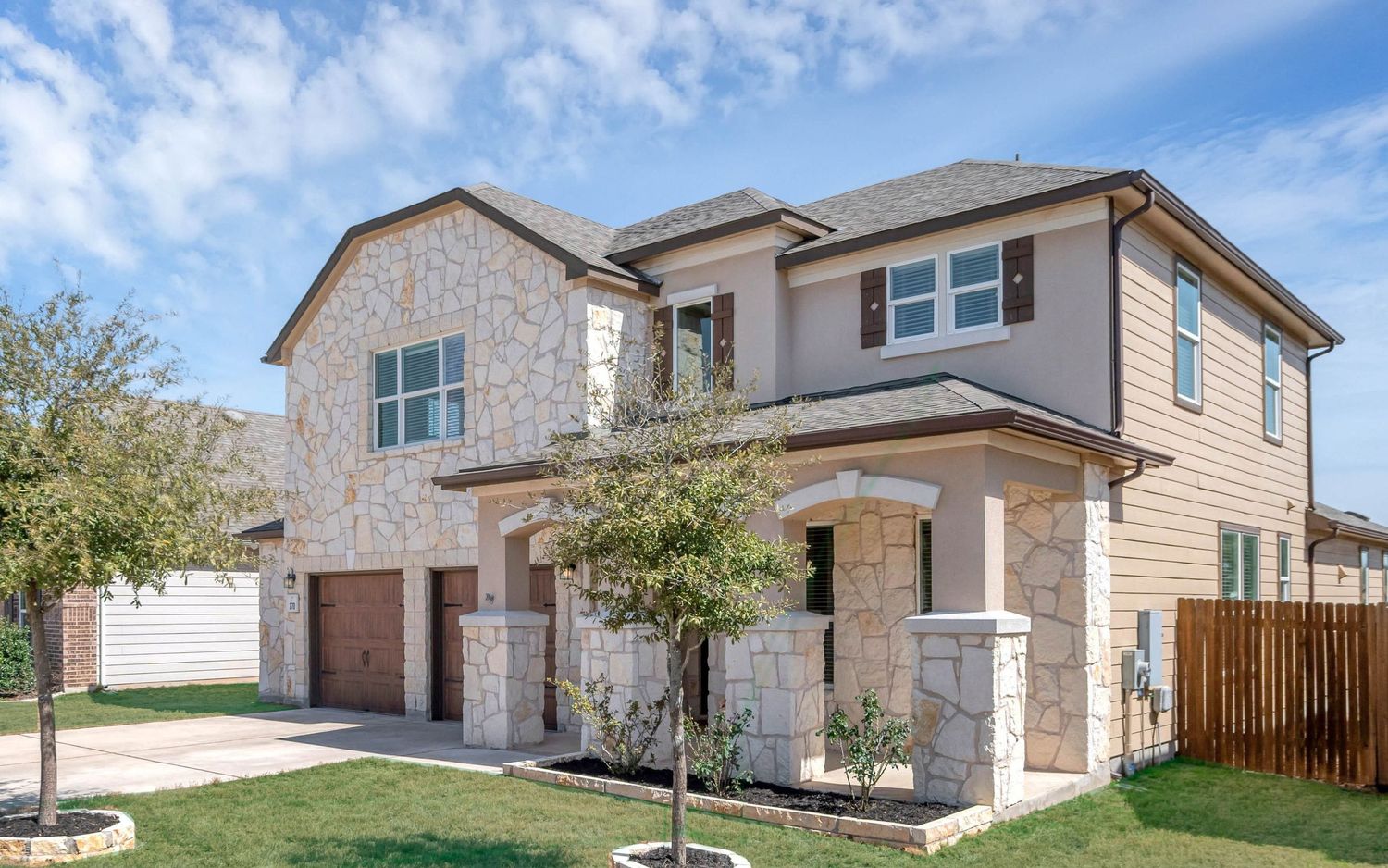 Two-story stone and siding home with a covered stone-pillar entry, brown garage door, young tree, and a wooden fence beside the yard.