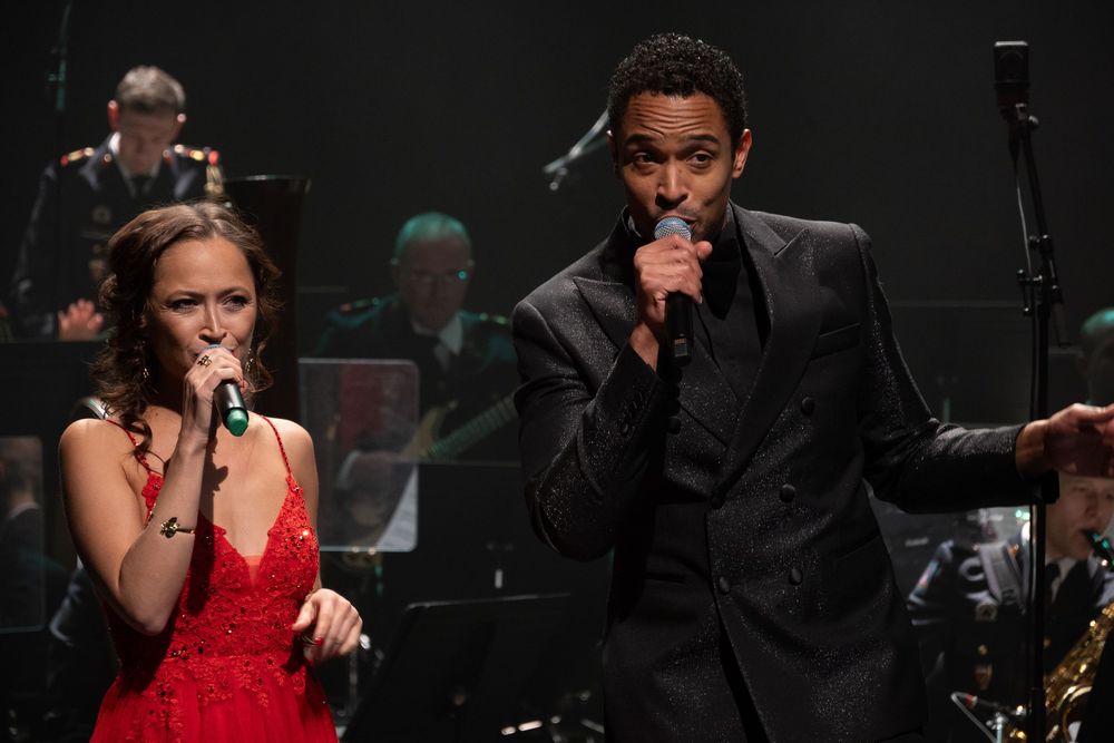 Chanteuse en robe rouge et chanteur en costume noir pailleté, micros en main, devant un orchestre en uniforme sur scène