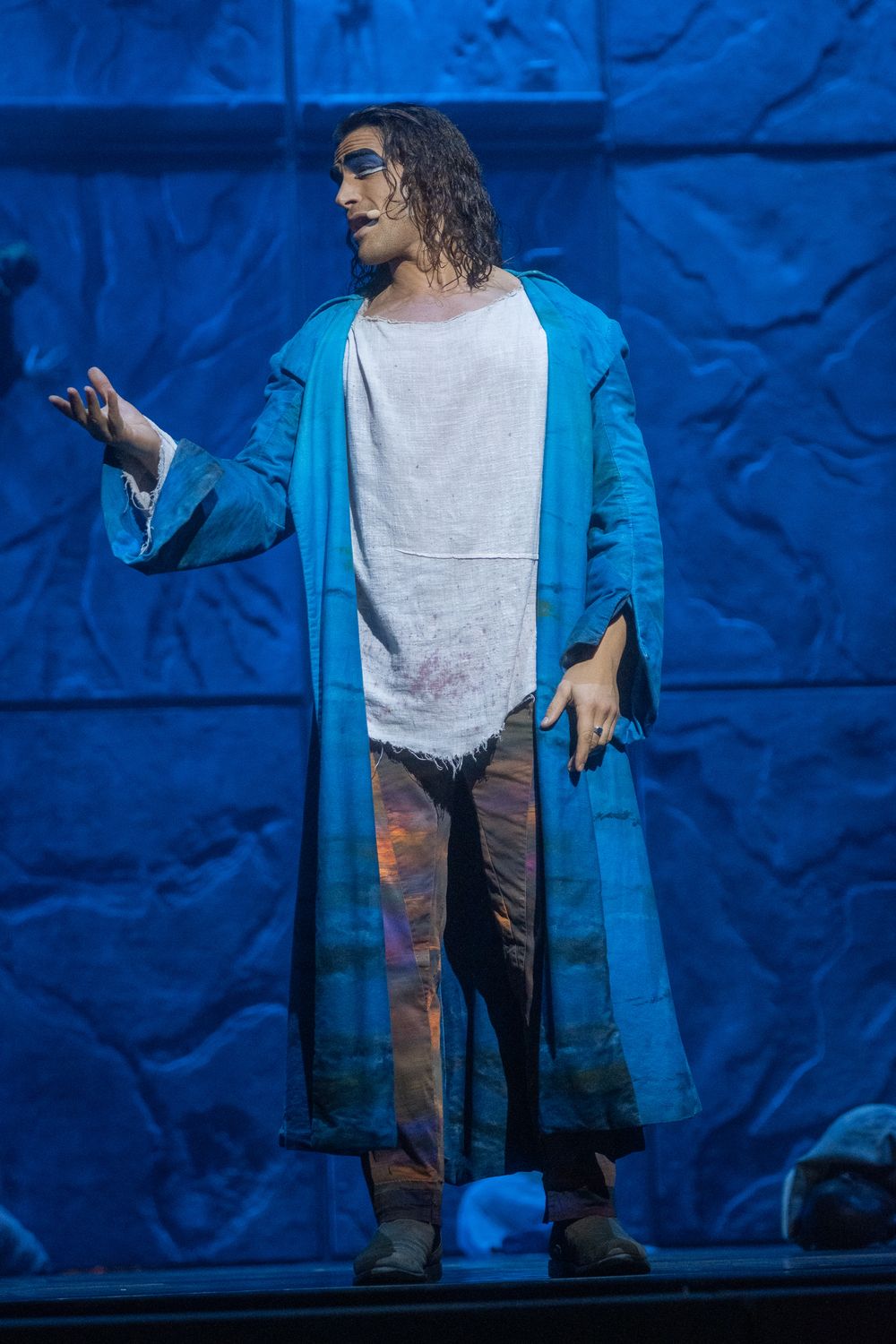 Chanteur aux longs cheveux bouclés sur scène, long manteau bleu turquoise sur tunique blanche, bras tendu, décor de pierre bleue